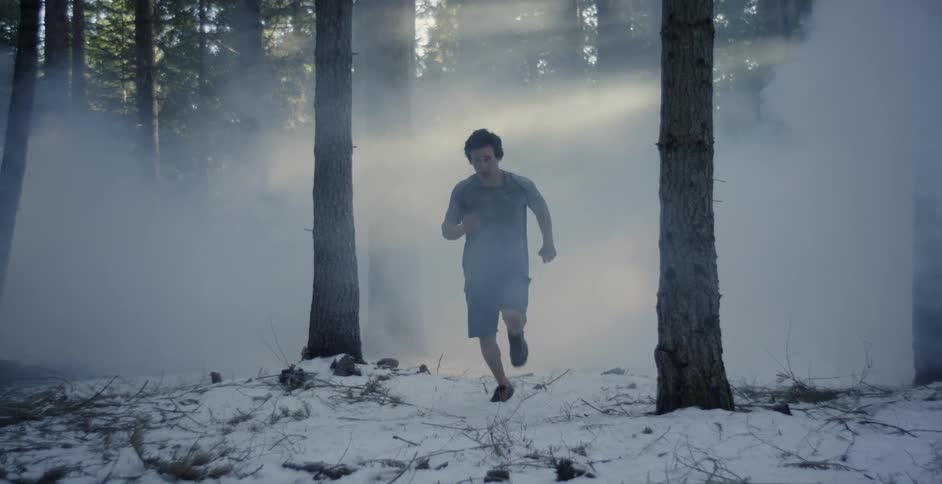A man in shorts is running across a snow-covered forest floor in daylight