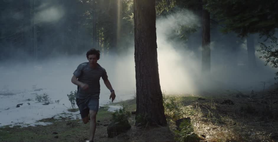 A man in shorts is running across a snow-covered forest floor on a foggy day