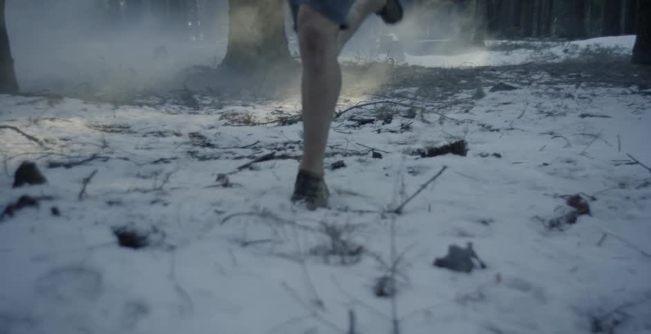 A man in shorts is running across a snow-covered forest floor in daylight