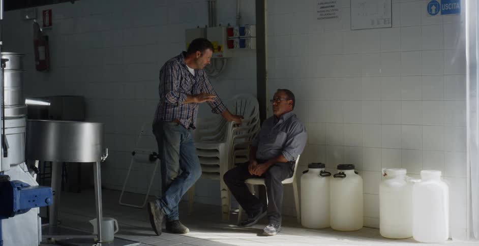 Two men sit and talk in a factory like setting