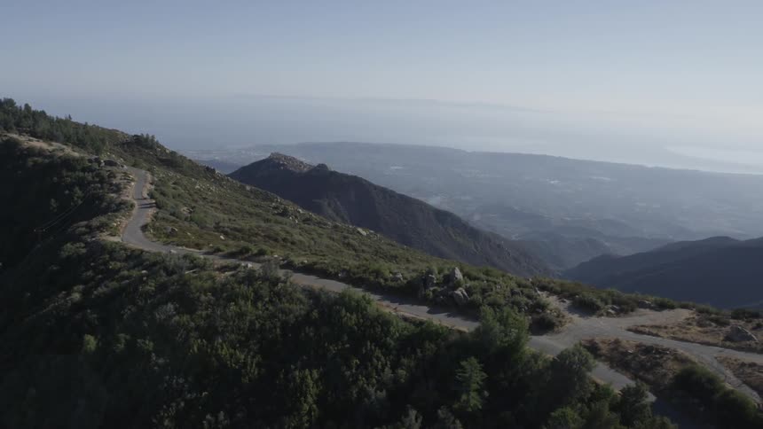 An aerial view of a narrow road winding through the mountains with other mountains in the background