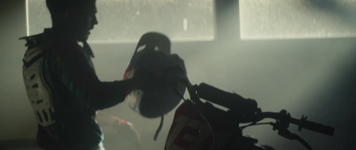 A man walks towards a dirt bike in a garage and puts on a helmet