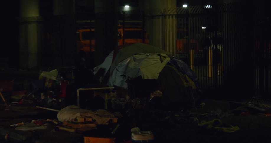 Trash and belongings litter a homeless camp in a city at night
