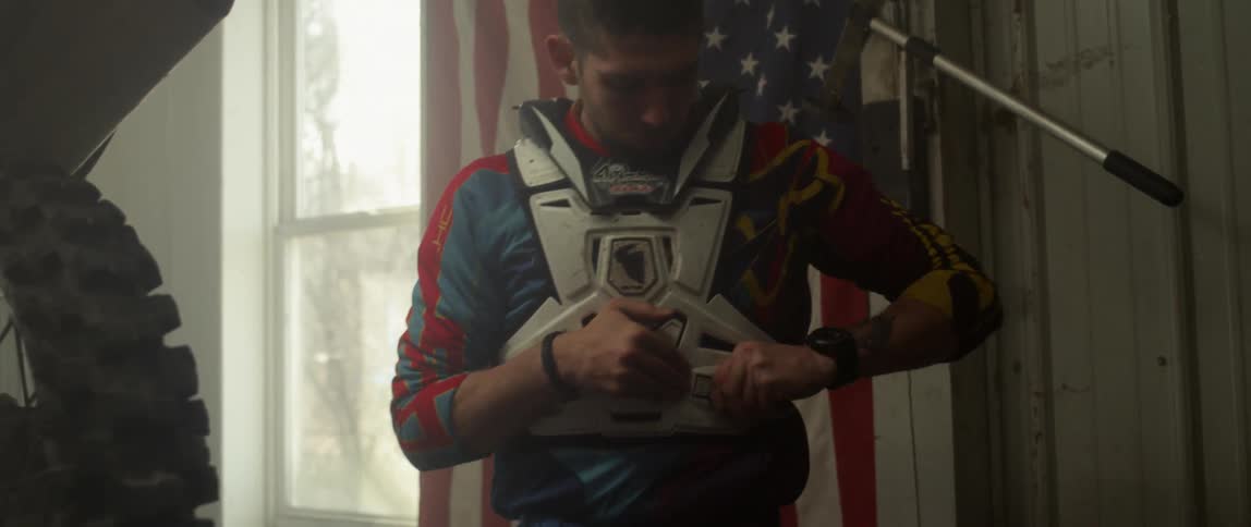 Man putting on protective racing gear indoors with an american flag hanging behind him