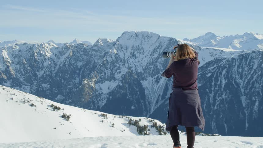 A woman is taking photos of a snow-covered mountain range on a sunny day
