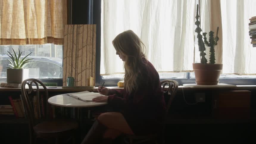A woman is thinking and writing inside at a table in daylight