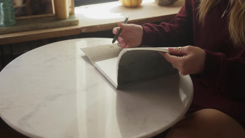 A woman sitting indoors at a table is taking out a writing notebook to write in
