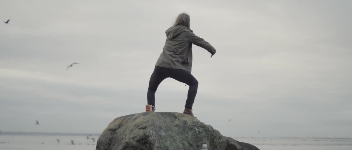 Woman waves her arms around standing on a large rock overlooking water