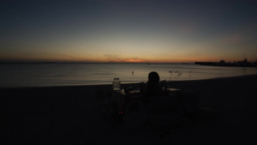 A woman is drinking a glass of wine as she sits at the shore at dusk