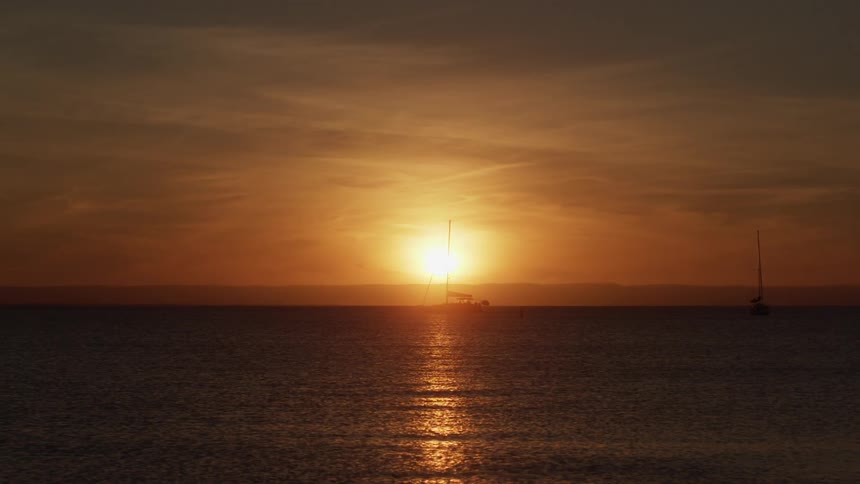 A sailboat on a large lake passes in front of the setting sun