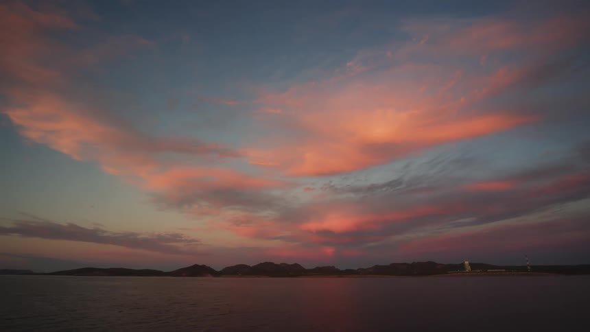 Colorful sunset occurring behind clouds and mountains in the distance before a large body of water