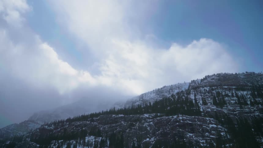 Clouds are streaming across snow-covered mountains in daylight