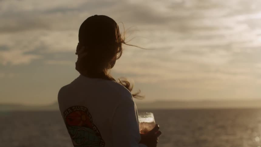 A woman is drinking a glass of wine as she watches the ocean at sunset