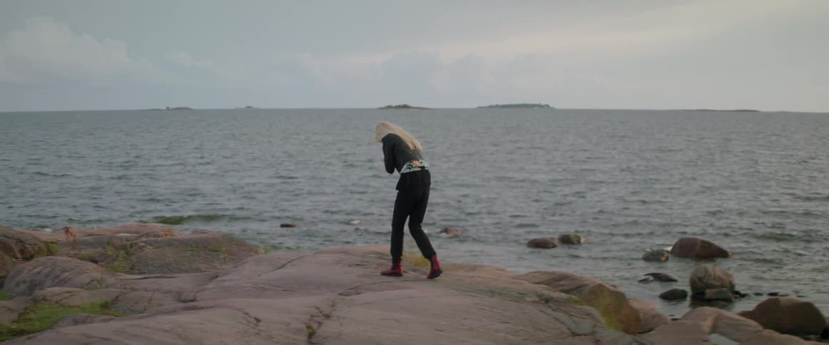 A distressed woman walks across a rocky shoreline and bends over in grief.