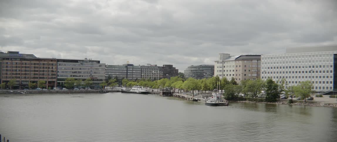 Lush green trees line an urban waterfront promenade under a cloudy summer sky.