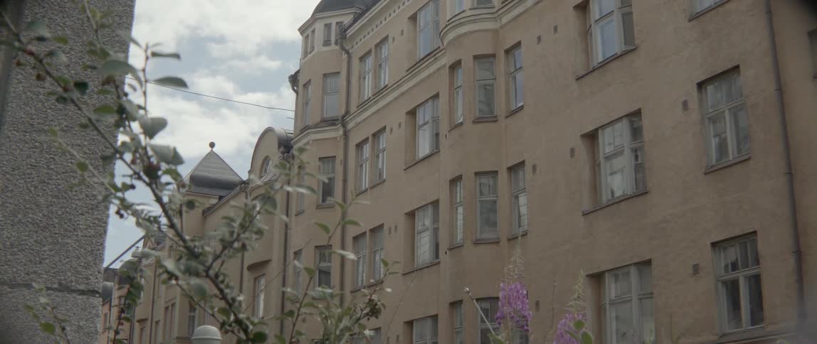 Purple flowers and green tree branches sway gently outside a classic beige apartment block.