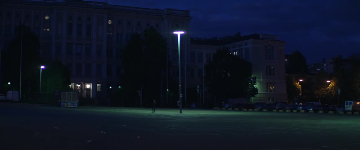 A lonely street lamp shines down on a dark open plaza near urban buildings.