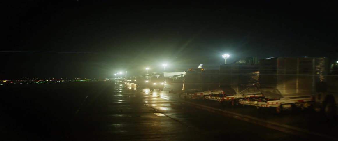Many trucks drive on the pavement near large lit up buildings at night