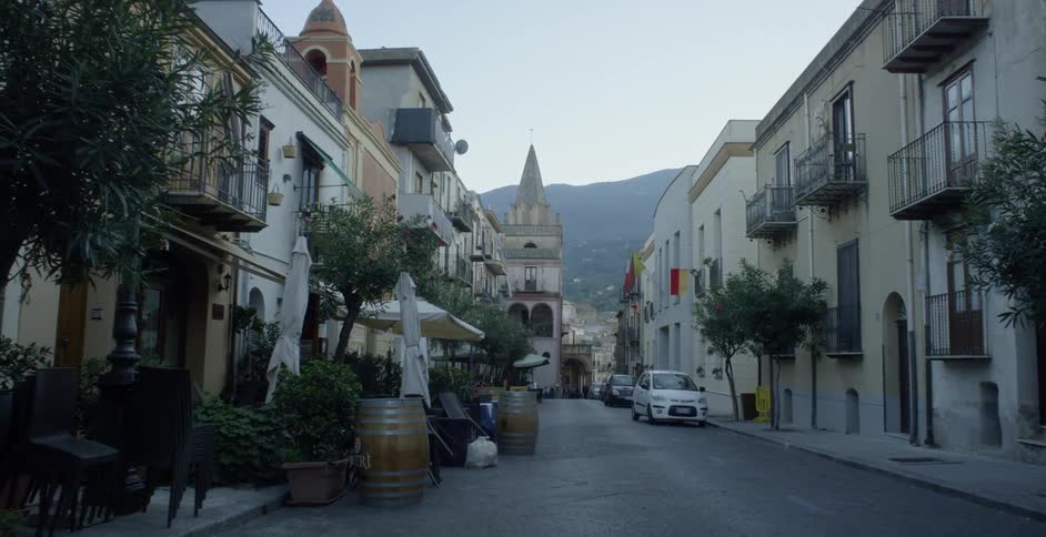 Cars are parked on a village road lined with shops and cafes