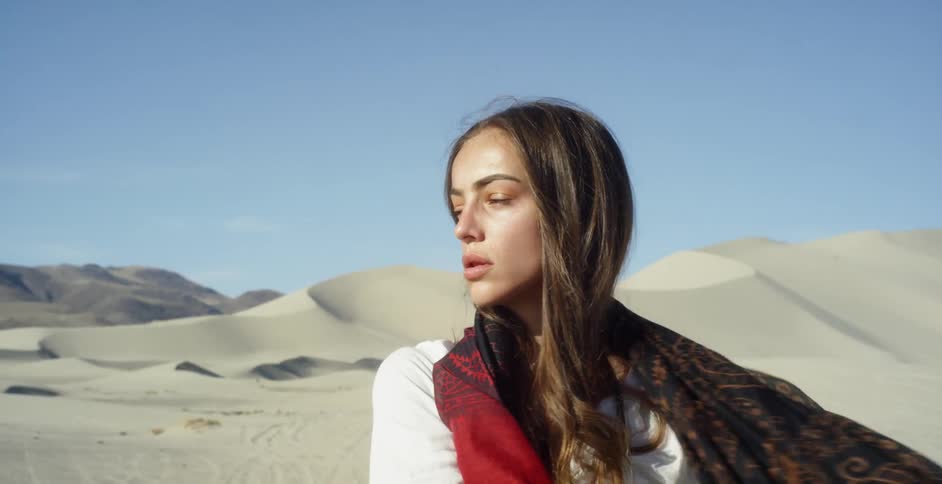 Long-haired brunette unwrapping a long scarf that was wrapped around her head and face in a sandy area on a clear day