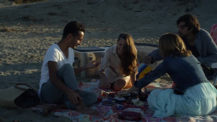 A group of friends sit on a blanket and eat as they picnic on the beach