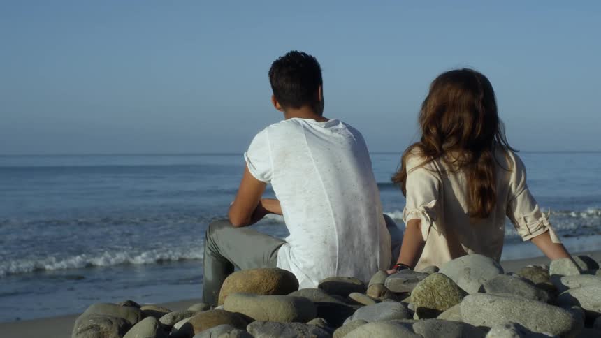 A man and woman sit on a rocky beach