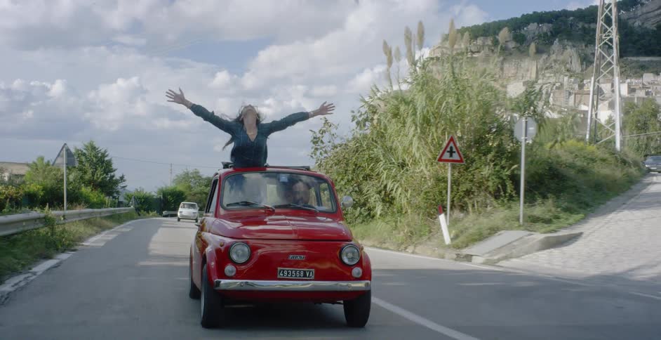 A woman stands arms open through the sunroof of a moving car