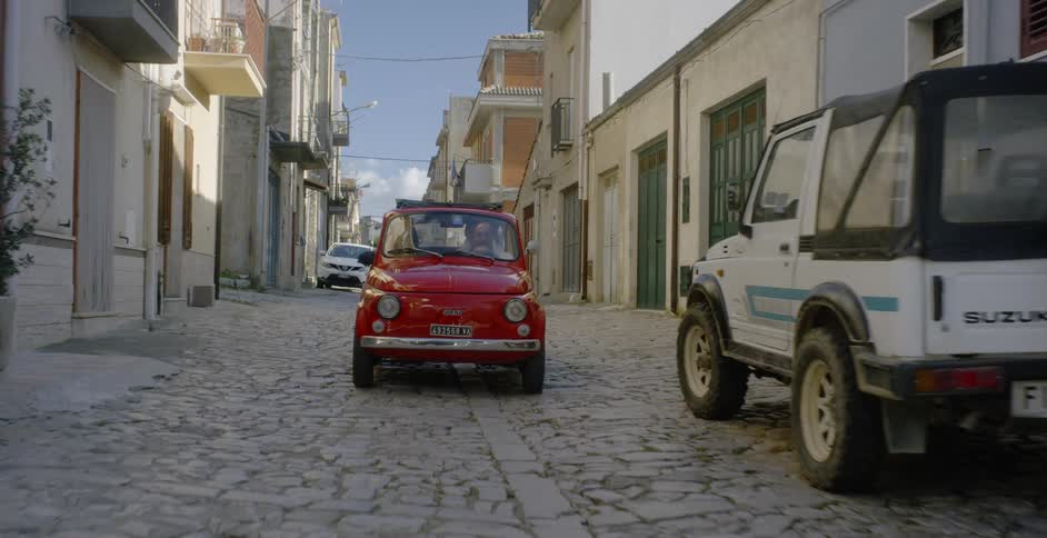Man and woman driving in small red car on cobblestone city road