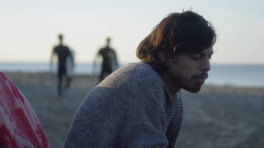 Man sits on the beach looking down as two people walk away