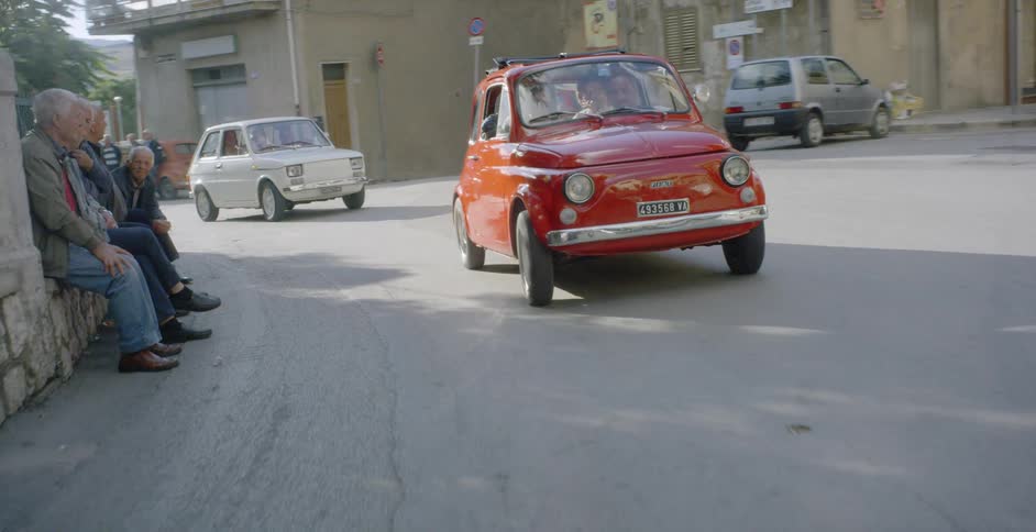 Cars drive past people sitting and standing along a village road