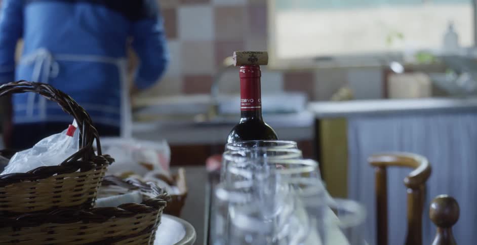 Wine bottle and glasses are lined up on a table while a person cooks