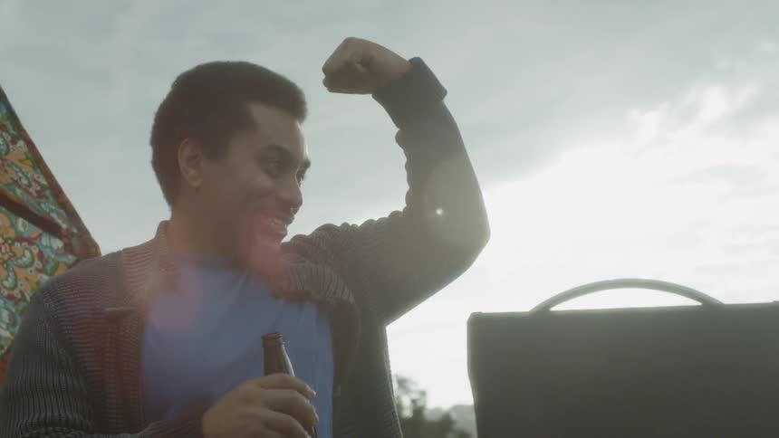 A man holds a beer bottle as he gestures with his arm and smiles