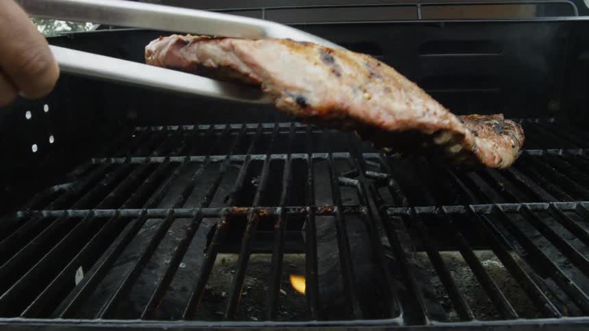 Tongs reach to turn a rack of ribs over as they cook on a grill