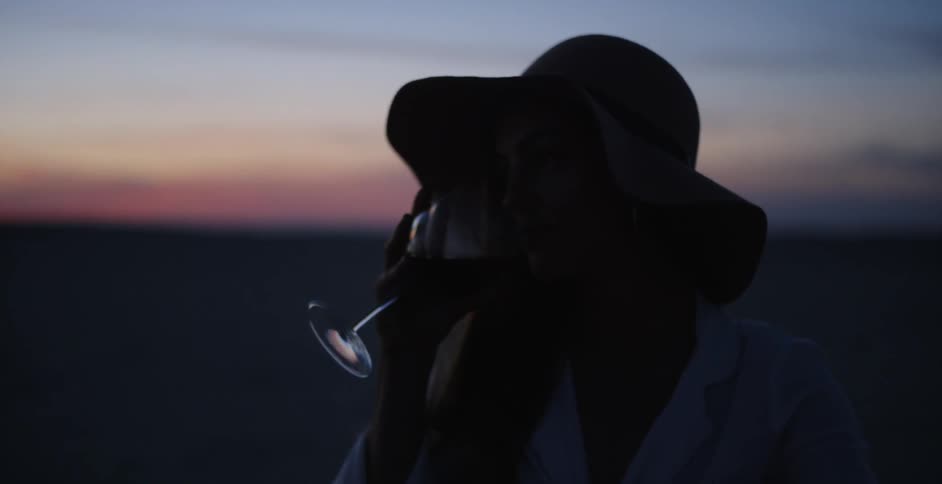 Woman in a hat drinks a glass of wine outside at dusk