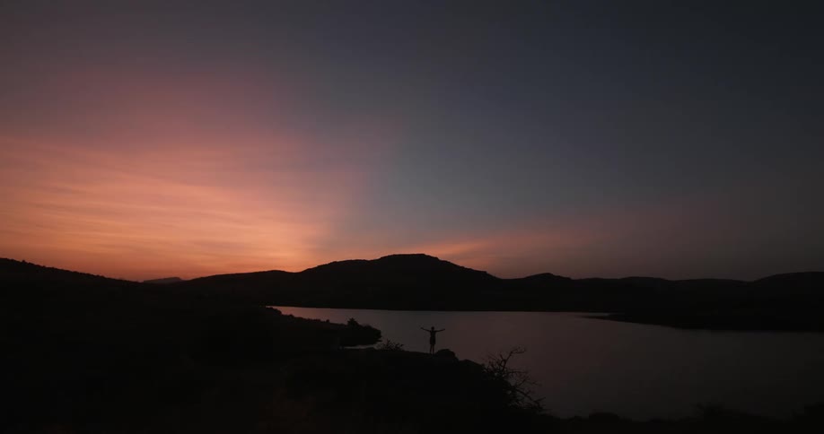 A person raises their arms by the lake at dawn