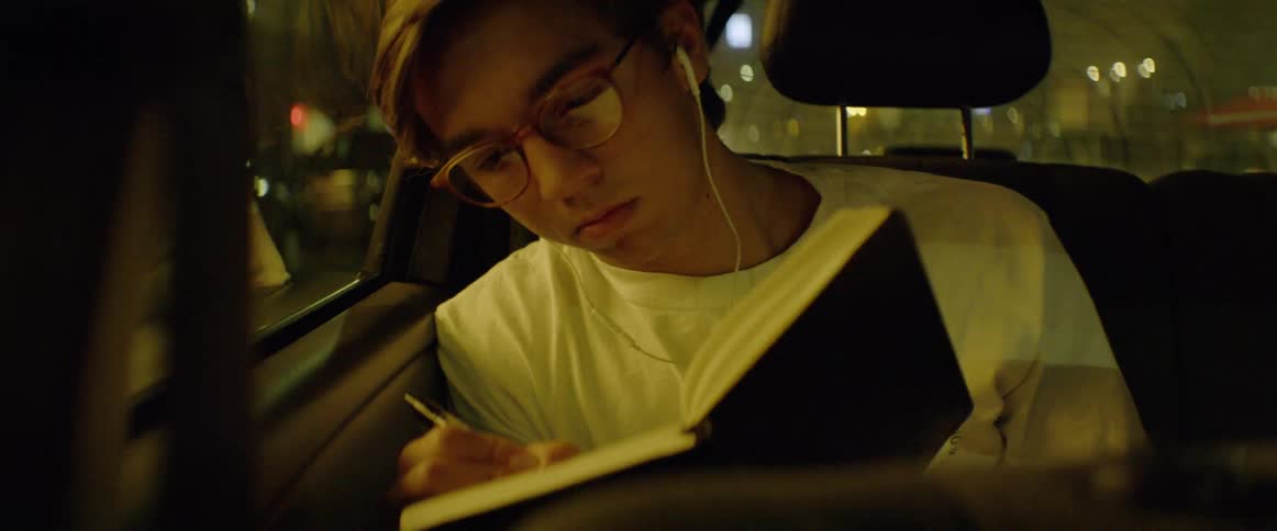 Boy rides in car listening to headphones and writing in journal at night