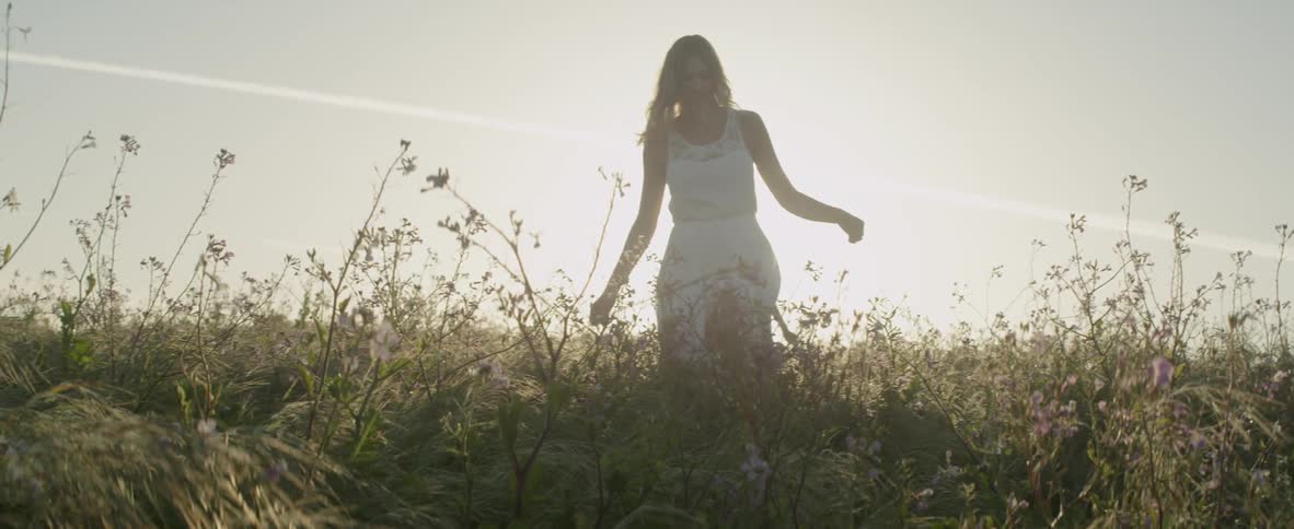 A woman is walking through a field of weeds and flowers on a sunny day