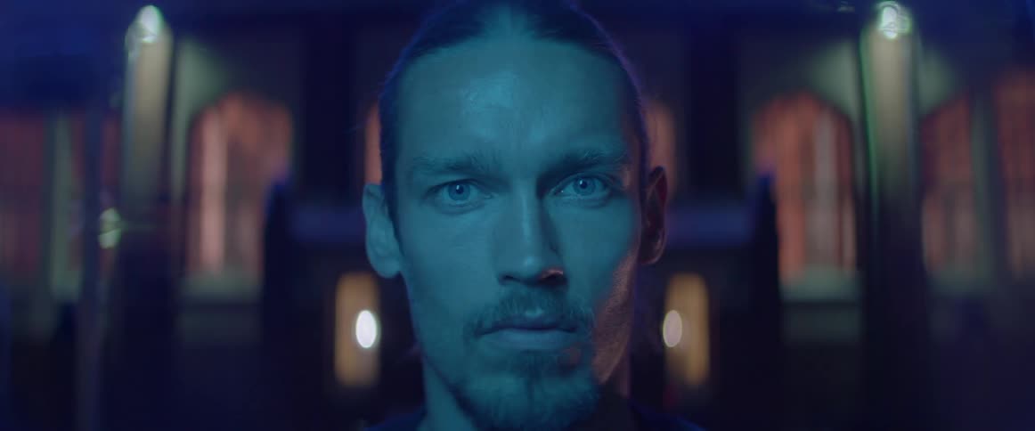 A serious young man stares directly into the camera under blue cinematic lighting.