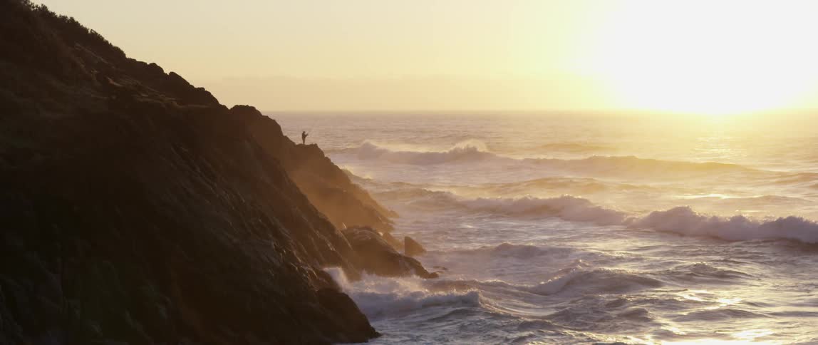 Man standing on a rocky cliff as the sun rises and the waves hit the rocks