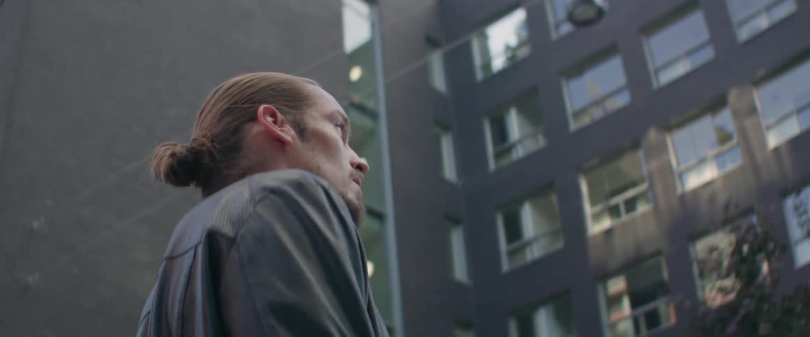 A man with a hair bun stands outside near contemporary city apartment blocks.