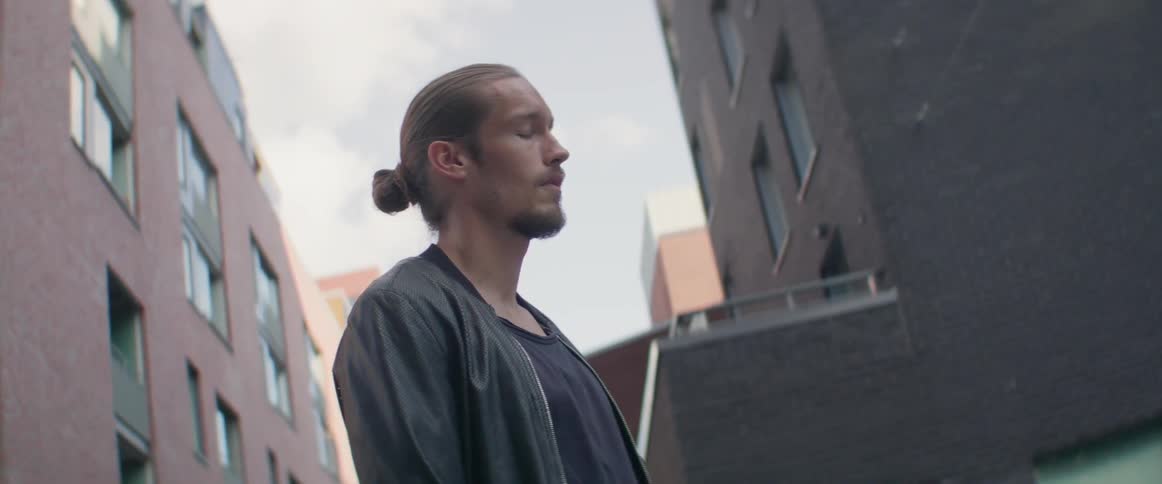 A young man with closed eyes stands peacefully outside modern residential apartment buildings.