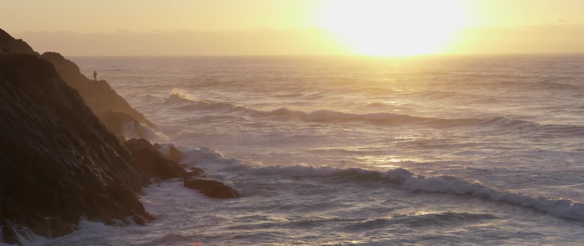 Man standing on a cliff as waves hit the rocks below and the sun rises