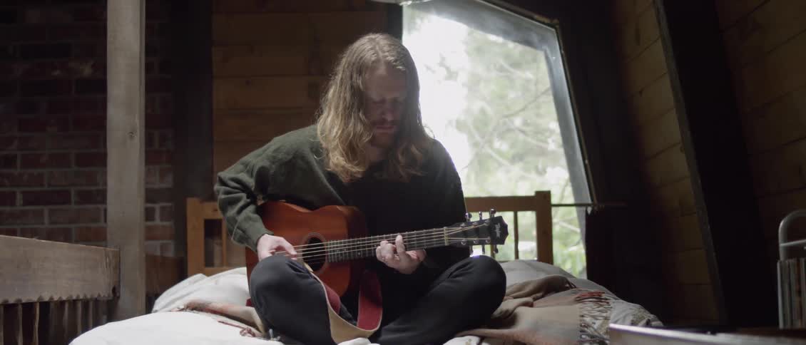 A man sits on the bed playing guitar and nodding his head