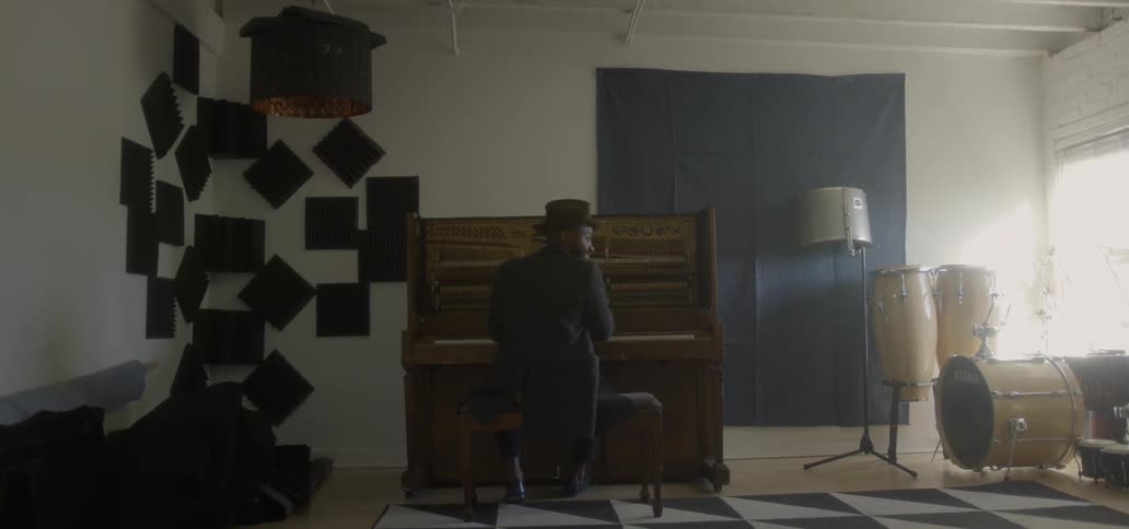 A man sits at a piano in a room while wearing a hat and coat