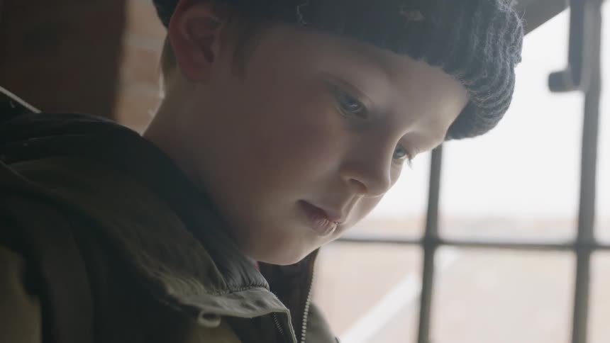 A boy is standing inside near a window concentrating on something