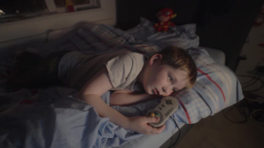 A boy is holding a video game controller as he lies in bed at night