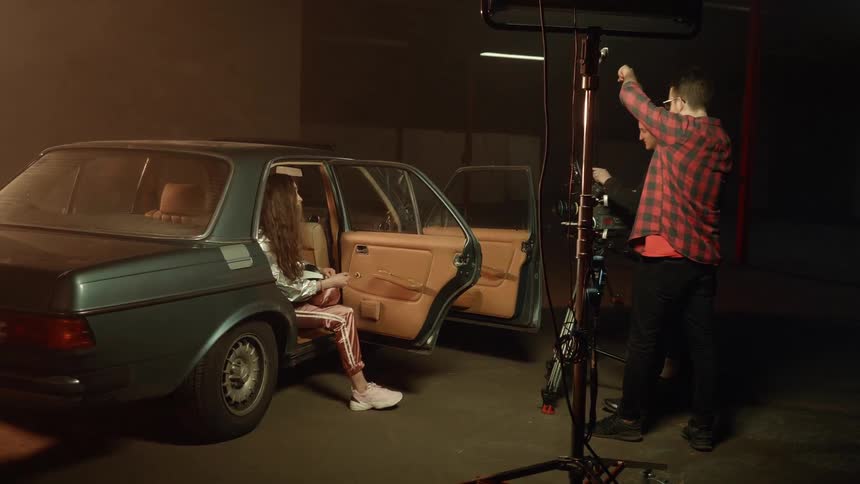 Woman sits in a car in a garage while a man films her