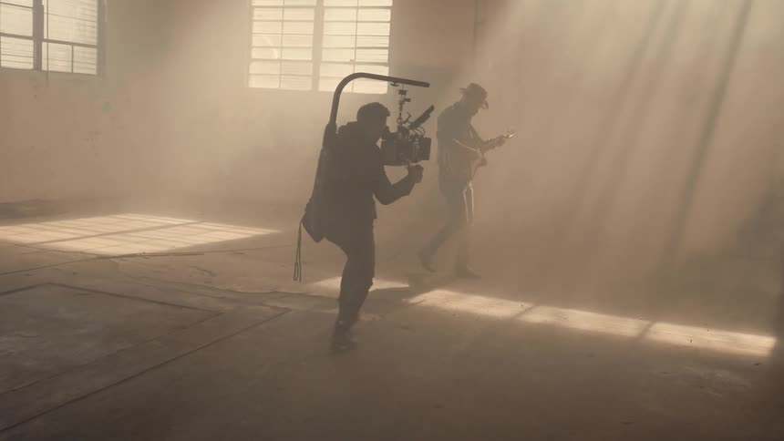 A man with a camera rig is filming another man playing guitar