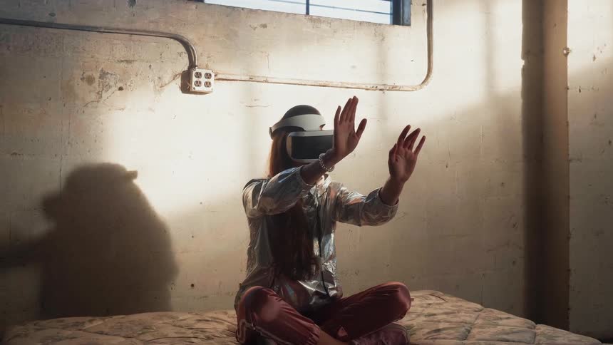 A woman wearing vr glasses sits on a mattress with her hands up