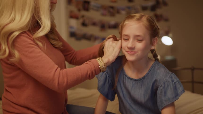 A woman braids a girl's hair while sitting on a bed